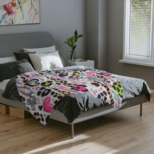 May include: A patterned blanket with a leopard print design, incorporating hearts, stars, and other graphics in pink, black, and green. The blanket is on a bed with a grey headboard and wooden flooring.