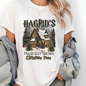 May include: White t-shirt with a graphic of a snowy cottage with Christmas lights and the text "Hagrid's Magically Grown Christmas Trees".