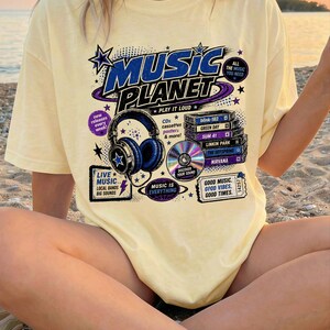 Puede incluir: Una camiseta amarilla clara con un diseño gráfico con las palabras "MUSIC PLANET" en azul y morado. El diseño incluye auriculares, CD y cintas de cassette. El texto adicional incluye "PLAY IT LOUD" y "MUSIC IS EVERYTHING".