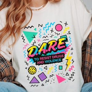 May include: Off-white t-shirt with a bold, colourful graphic. The design prominently displays "D.A.R.E." in a multi-coloured font, and the text "TO RESIST DRUGS AND VIOLENCE." Geometric shapes and lightning bolts complete the design.