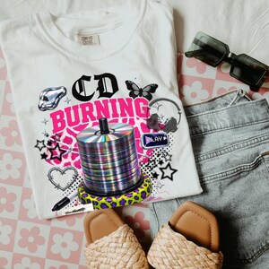 May include: White Comfort Colors t-shirt with a graphic design that says "CD BURNING" in black and pink. The design includes a stack of CDs, a butterfly, stars, and other elements. The shirt is paired with sunglasses, denim shorts, and sandals.