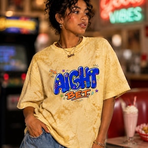 May include: A yellow acid-wash t-shirt featuring a graphic print with the text AIGHT BET in large blue bubble letters with orange outlines. The design includes small colorful stars, swirls, and an orange arrow. The shirt has a crew neckline.