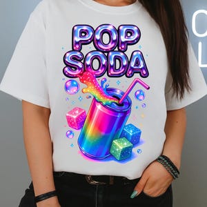 Puede incluir: Camiseta blanca con un gráfico vibrante de una lata de refresco arcoíris, burbujas y cubos brillantes. El texto "POP SODA" está en una fuente brillante y multicolor. Un diseño divertido y colorido.