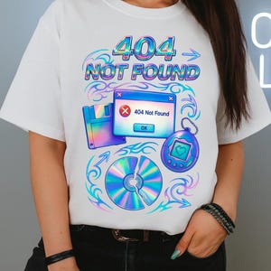 Op de afbeelding: Wit T-shirt met een kleurrijk grafisch ontwerp. Het ontwerp bevat de tekst "404 NOT FOUND" en retro computergerelateerde iconen. Het bevat een diskette, een kapotte cd en een Tamagotchi-stijl sleutelhanger.