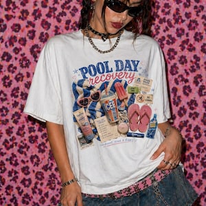 Pode incluir: Camiseta branca com um gráfico que diz "POOL DAY recovery" com óculos de sol, sorvete e outros itens de verão. A modelo está usando uma saia jeans e acessórios. O fundo é uma estampa de leopardo rosa.