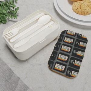 May include: A white lunch box with a fork, spoon, and knife inside. Next to it is a coaster with a pattern of VHS tapes, each labeled "BE KIND REWIND" in white text with a rainbow stripe.
