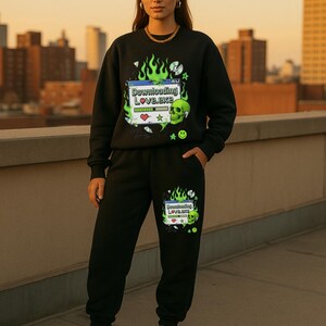 Può includere: Completo nero composto da felpa e pantaloni della tuta con un design grafico. Il design include il testo "Downloading Love.exe", un teschio e fiamme. L'outfit è indossato con scarpe da ginnastica bianche.