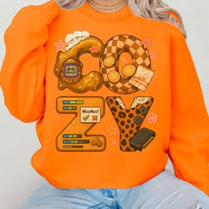 May include: Bright orange sweatshirt with the word "COZY" in a playful, illustrated design. The "C" is a brown, textured shape with a small screen. The "O" is a checkered pattern. The "Z" has a game-like interface. The "Y" is leopard print.