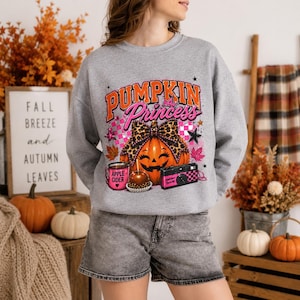 May include: A heather gray crewneck sweatshirt featuring a graphic print with the text PUMPKIN Princess, a jack-o'-lantern wearing a leopard print bow, a pink mug labeled Apple Cider, a caramel apple, and a pink camera with a checkered pattern.