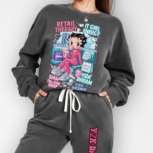 Può includere: Completo composto da felpa e pantaloni della tuta grigio scuro. La felpa presenta una grafica di un personaggio dei cartoni animati con la scritta "RETAIL THERAPY IT GIRL ENERGY" e "Y2K DREAM". I pantaloni della tuta hanno "Y2K Dream" stampato sulla gamba.