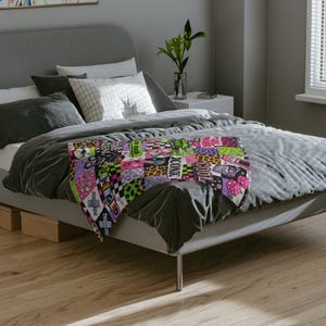 Könnte beinhalten: Eine farbenfrohe Patchwork-Decke mit verschiedenen Mustern, darunter Leopardenmuster und Schachbrettmuster, sowie Text. Die Decke in Pink, Grün, Lila und Schwarz ist über ein Bett mit grauer Bettwäsche drapiert.