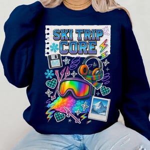 Op de afbeelding: Marineblauwe sweatshirt met een kleurrijk grafisch ontwerp. Het ontwerp bevat de tekst "SKI TRIP CORE", een skibril, een koptelefoon, een berglandschap en andere winterse elementen. De sweatshirt heeft een ronde hals.