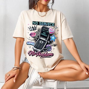 Może przedstawiać: Beżowa koszulka z nadrukiem graficznym przedstawiającym telefon z klapką, napisy "NO SERVICE" i "No Problem" oraz inne grafiki w stylu retro. Projekt zawiera koronę, usta i serce.
