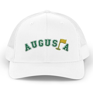 May include: A white trucker cap featuring an embroidered design on the front. The text reads AUGUS with a yellow golf flag replacing the letter T, followed by the letter A. The embroidery is in dark green with a white outline and yellow details.