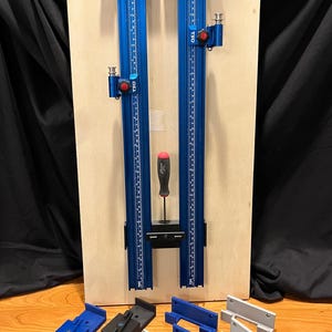May include: Two blue metal TSO parallel guide rails with measurement markings are mounted on a wooden board. A screwdriver is placed between the rails. Several black, blue, and grey accessories are in the foreground.