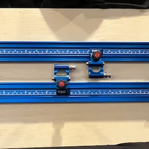 May include: Two blue TSO parallel guide rails with measurement markings, black brackets, and blue and green adjustment wheels. The rails are mounted on a light wood surface.