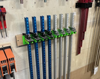 May include: A collection of woodworking clamps in various colors, mounted on a light wood wall. The clamps include blue and silver bar clamps, along with red and orange clamps. The clamps are organized and ready for use in a workshop setting.