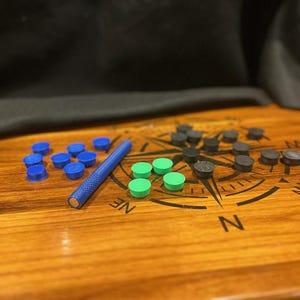 May include: A set of small round game pieces in blue, green, and black, accompanied by a blue cylindrical tool with a textured grip. These items are arranged on a wooden surface featuring a printed compass rose design with cardinal directions.
