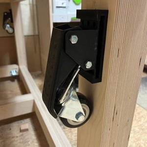 May include: A black caster wheel attached to a wooden workbench leg. The caster has a silver wheel and is secured with bolts. The workbench is made of light-colored wood.