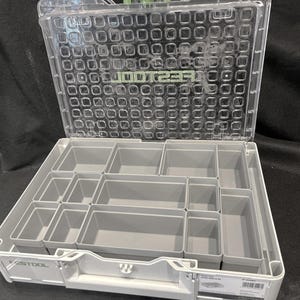 Puede incluir: Un organizador de plástico gris y transparente con múltiples compartimentos. La tapa transparente está abierta, revelando contenedores de almacenamiento grises de varios tamaños. El nombre de la marca es visible en la parte delantera. El organizador está diseñado para herramientas o piezas pequeñas.