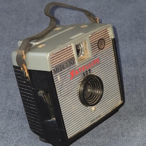 May include: Vintage Mercury Satellite 127 camera. The camera is gray and black with a silver face and a black lens. The words "Mercury Satellite 127" are printed on the front in red. The camera has a black strap and is marked "Made in USA".