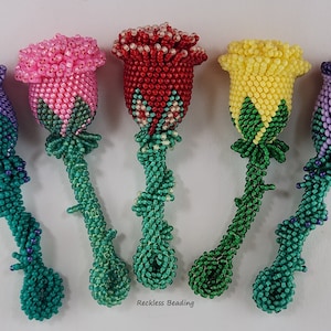 Könnte beinhalten: Fünf Perlenrosen-Stiele in verschiedenen Farben, darunter Lila, Rosa, Rot, Gelb und Lavendel. Die Stiele sind grün und haben ein gedrehtes Design. Die Rosen bestehen alle aus kleinen Perlen und haben ein realistisches Aussehen. Der Text "Reckless Beading" ist am unteren Rand des Bildes sichtbar.