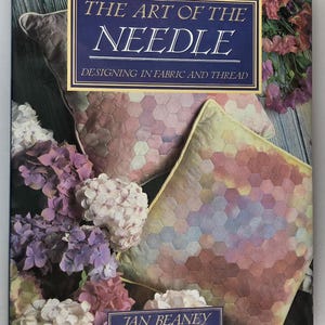 May include: A hardcover book titled The Art of the Needle: Designing in Fabric and Thread by Jan Beaney. The cover features two square pillows with a multicolored hexagonal patchwork pattern, surrounded by clusters of purple and white hydrangea flowers.