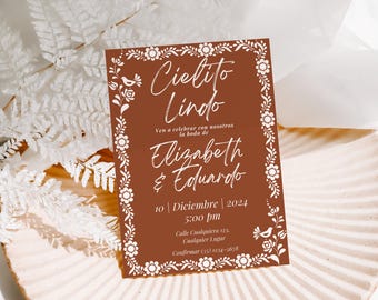 May include: A brown wedding invitation with a white floral border. The text reads "Cielito Lindo" and includes the names "Elizabeth & Eduardo", the date "10 | Diciembre | 2024", the time "5:00 pm", the address "Calle Cualquiera 123. Cualquier Lugar", and the phone number "(55) 1234-5678".