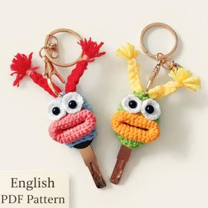 May include: Two handmade crochet keychains, one blue and red, the other green and orange, each with large googly eyes and tassel accents. The keychains are attached to gold-colored key rings. The image includes a sign that says "English PDF Pattern".
