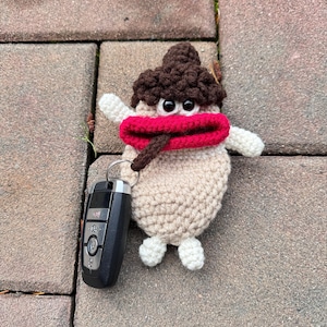 May include: A crocheted key chain shaped like a cartoon character with brown hair, a red mouth, and large black eyes. The keychain is attached to a black car key fob.