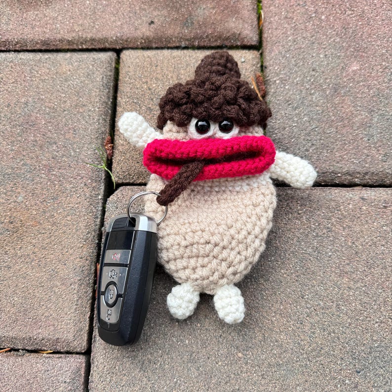 May include: A crocheted key chain shaped like a cartoon character with brown hair, a red mouth, and large black eyes. The keychain is attached to a black car key fob.