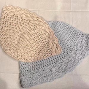 May include: Two crocheted items, one beige and one light blue, are displayed. Both have a scalloped edge and a textured, open weave pattern. The items appear to be handmade, possibly dishcloths or decorative pieces.