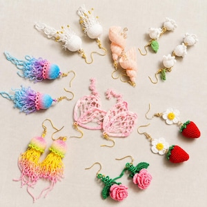 May include: A collection of handmade crochet earrings featuring diverse designs including jellyfish with colorful fringe, white floral sprigs, pink heart-shaped lace, red strawberries with white daisies, and pink roses with green leaves on gold-tone hooks.