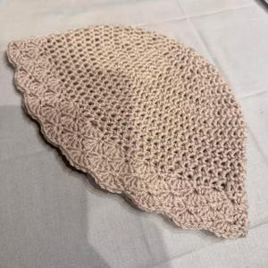 May include: A crocheted hat in a light beige color. The hat has a scalloped edge and a loose, open weave design. The hat is handmade and appears to be made of a soft yarn.
