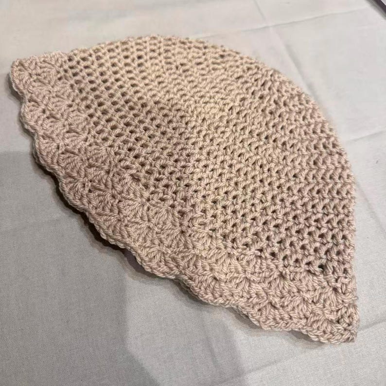 May include: A crocheted hat in a light beige color. The hat has a scalloped edge and a loose, open weave design. The hat is handmade and appears to be made of a soft yarn.