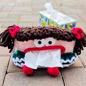 May include: A crocheted tissue box cover shaped like a girl with brown hair, pink skin, and big eyes. The cover has a green and white dress and red shoes.