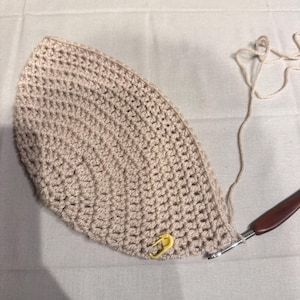 May include: A beige crocheted hat in progress, with a yellow stitch marker and a crochet hook. The hat is made with a textured stitch, and the yarn is a matching beige color. The project is on a white surface.