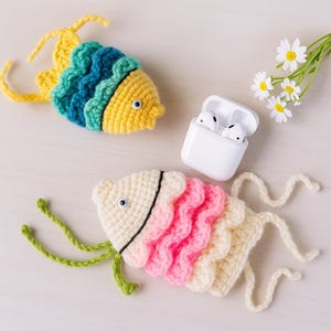 May include: Two crochet fish-shaped AirPod cases in yellow, blue, and pink. One case is yellow with blue and green accents, the other is white with pink and cream accents. A white AirPod case with two white earbuds is also present.