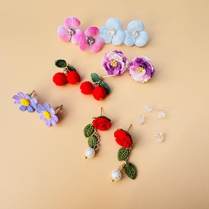 May include: A collection of handmade earrings in various floral and fruit designs. Includes pink, blue, and purple flower earrings, red cherry earrings, and red rose earrings with pearl accents. Clear earring backs are also visible.