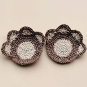 May include: Two brown and white crocheted coasters shaped like paw prints.