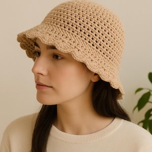 May include: A beige crochet bucket hat with a scalloped brim. The hat has an open weave design and a small brim. The hat is made of a soft, natural-colored yarn. The hat is a stylish accessory for sun protection.