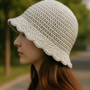 May include: A light beige crochet bucket hat with a scalloped brim. The hat is made of a textured yarn and has a small, downward-sloping brim. The hat is a neutral color and is suitable for various outfits.