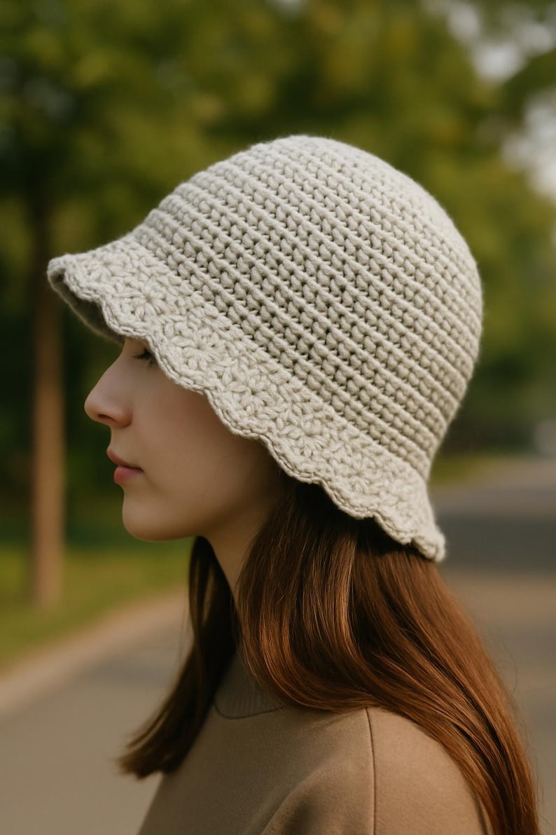 May include: A light beige crochet bucket hat with a scalloped brim. The hat is made of a textured yarn and has a small, downward-sloping brim. The hat is a neutral color and is suitable for various outfits.
