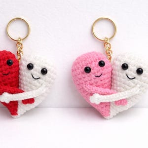Könnte beinhalten: Zwei gehäkelte Herz-Schlüsselanhänger, einer rot und weiß, der andere rosa und weiß, jeweils mit einem lächelnden Gesicht und einem Umarmungsdesign. Die Schlüsselanhänger haben goldfarbene Metallringe.
