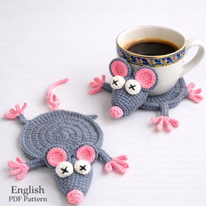 May include: Crocheted gray mouse-shaped coasters with pink ears, noses, and feet. The coasters have white eyes with black crosses and are paired with a white coffee cup. The image includes the text "English PDF Pattern".