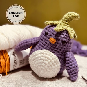 May include: A crocheted purple penguin with a white belly and a green crocheted leaf on its head. The penguin is sitting on a white surface with yarn in the background.