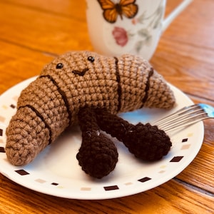 May include: A crocheted brown croissant with a smiling face and two brown legs on a white plate with a black and white geometric pattern.