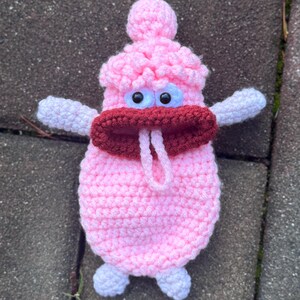 May include: A pink crocheted plush toy with large white eyes, a red mouth, and white arms. The toy has a small pink pom-pom on top of its head.
