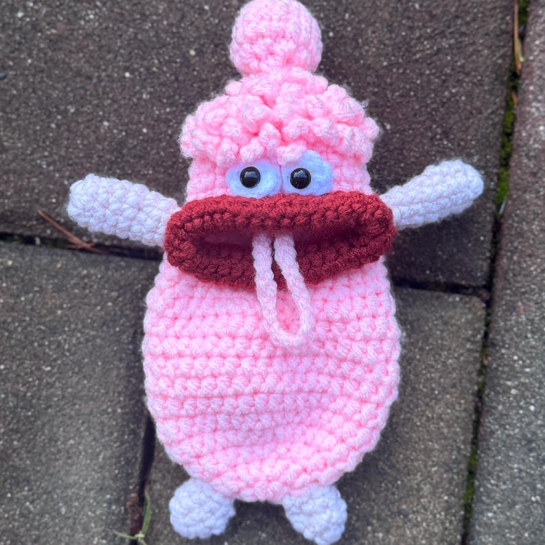 May include: A pink crocheted plush toy with large white eyes, a red mouth, and white arms. The toy has a small pink pom-pom on top of its head.