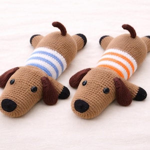 May include: Two crocheted dachshund dog toys. The dogs are brown with black eyes, noses, and paws. One dog wears a blue and white striped shirt, and the other wears an orange and white striped shirt. The toys are lying on a white surface.